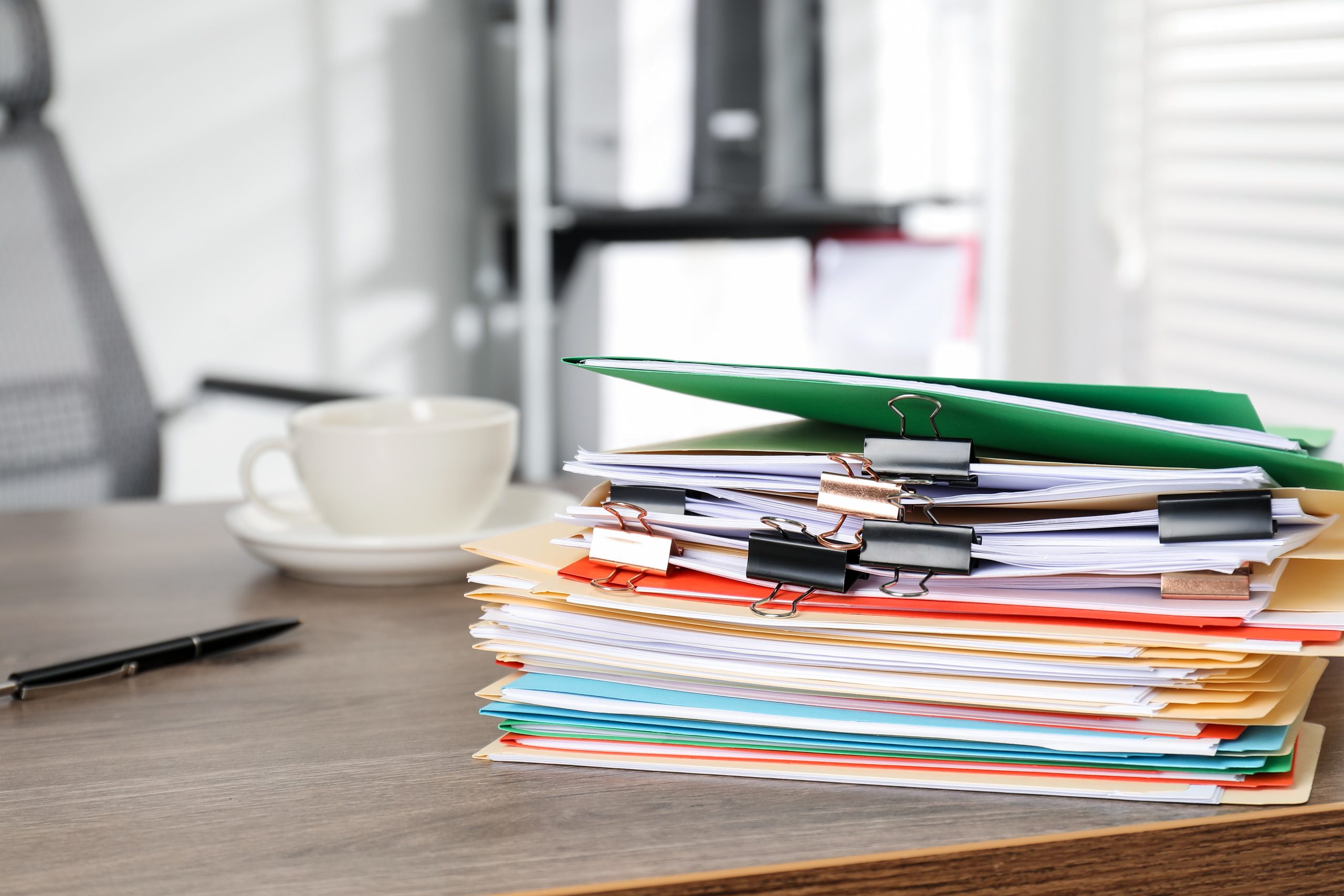 Towering stack of paperwork on a cluttered desk, symbolizing how “running lean” quietly shifts costs into overload, admin drag, and hidden operational risk.